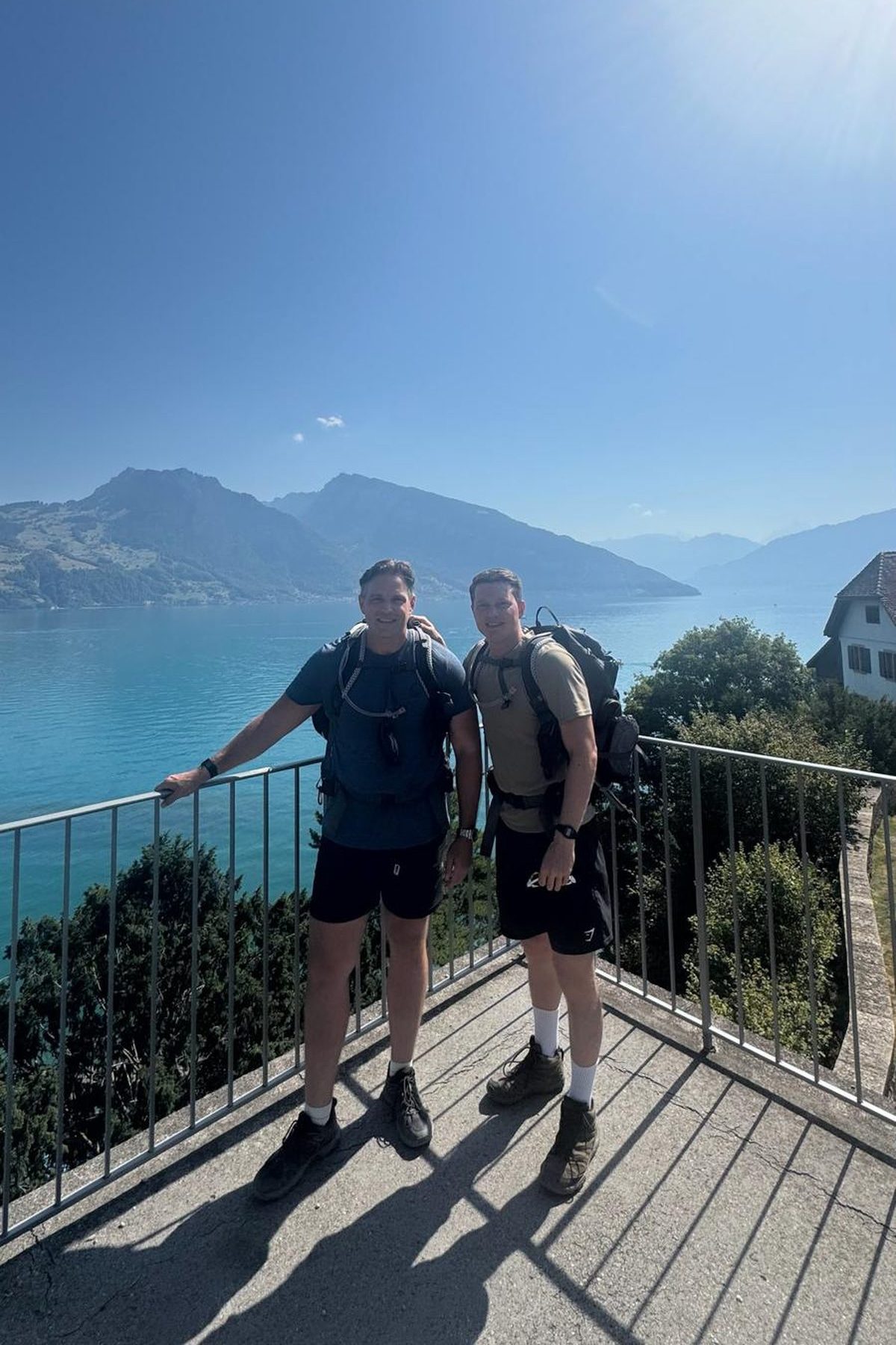 The two Wine Junkies in hiking gear with backpacks, standing at a railing above the turquoise water of Lake Thun